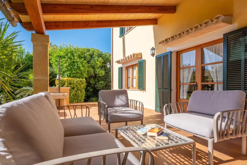 An outdoor seating area beside a house, with a glass-topped table and leafy trees.