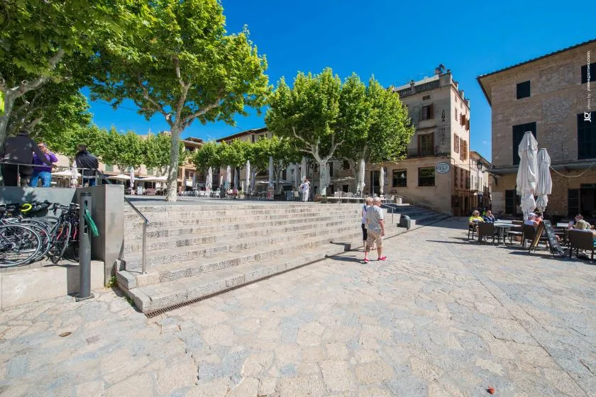 A sunny town square with broad stone steps, leafy trees, and outdoor café seating.