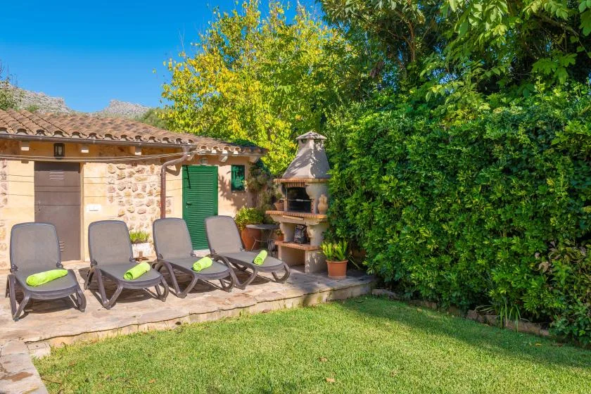 A garden patio with reclining loungers, a stone barbecue, and leafy greenery.