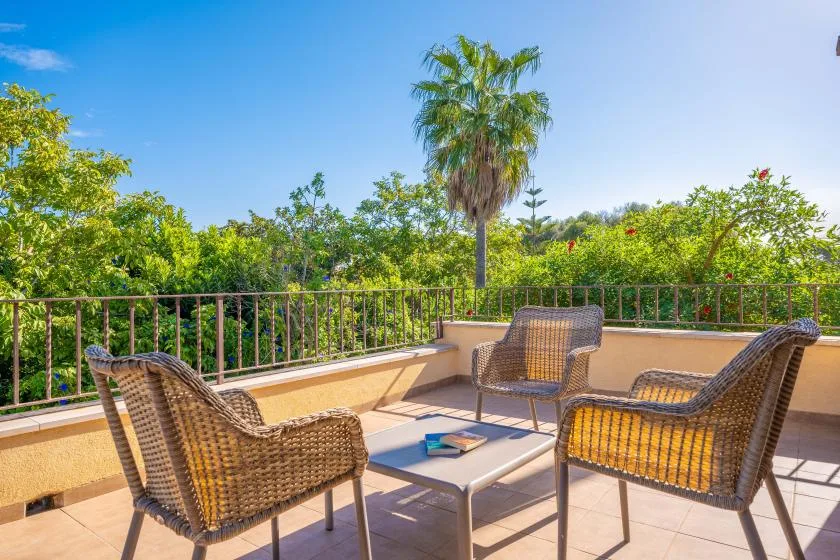 A sunlit outdoor patio has woven seating around a low table, with greenery beyond the railing.