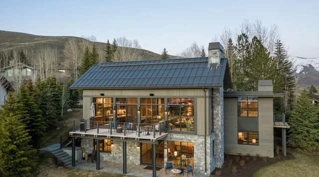 Una casa moderna con techo oscuro inclinado, grandes ventanas y una terraza elevada.
