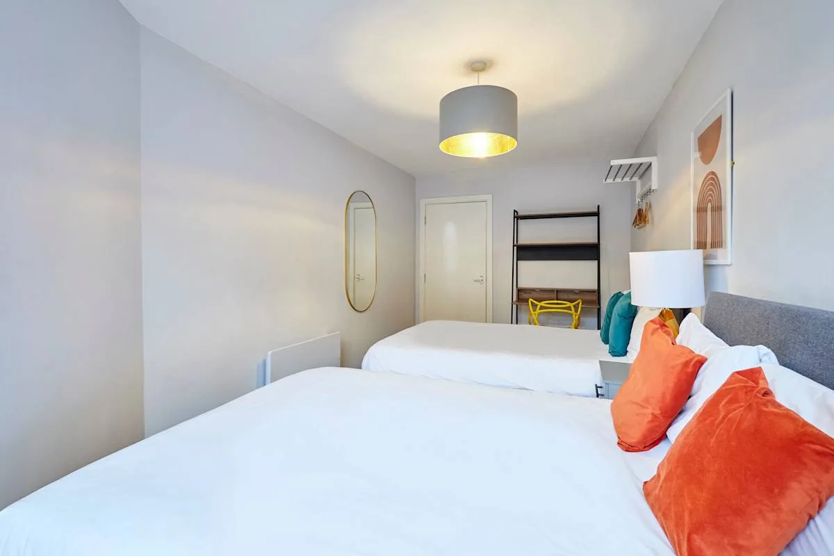 A bright bedroom with white bedding, colorful pillows, and a bedside lamp.