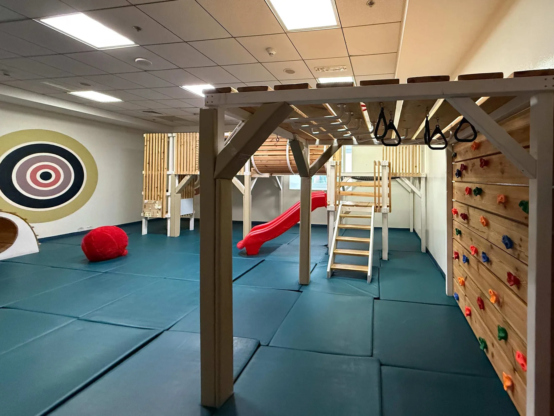 Zona de juegos interior con suelo acolchado, un tobogán y estructuras para trepar.