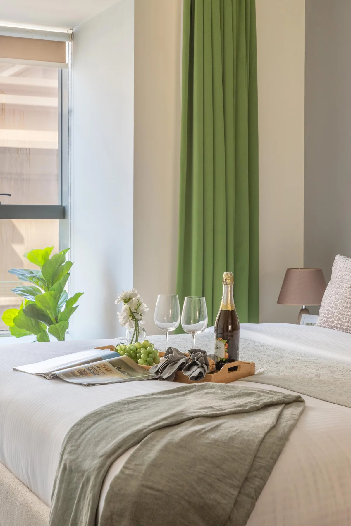 Una cama bien tendida está junto a una ventana con cortinas verdes y una pequeña bandeja.