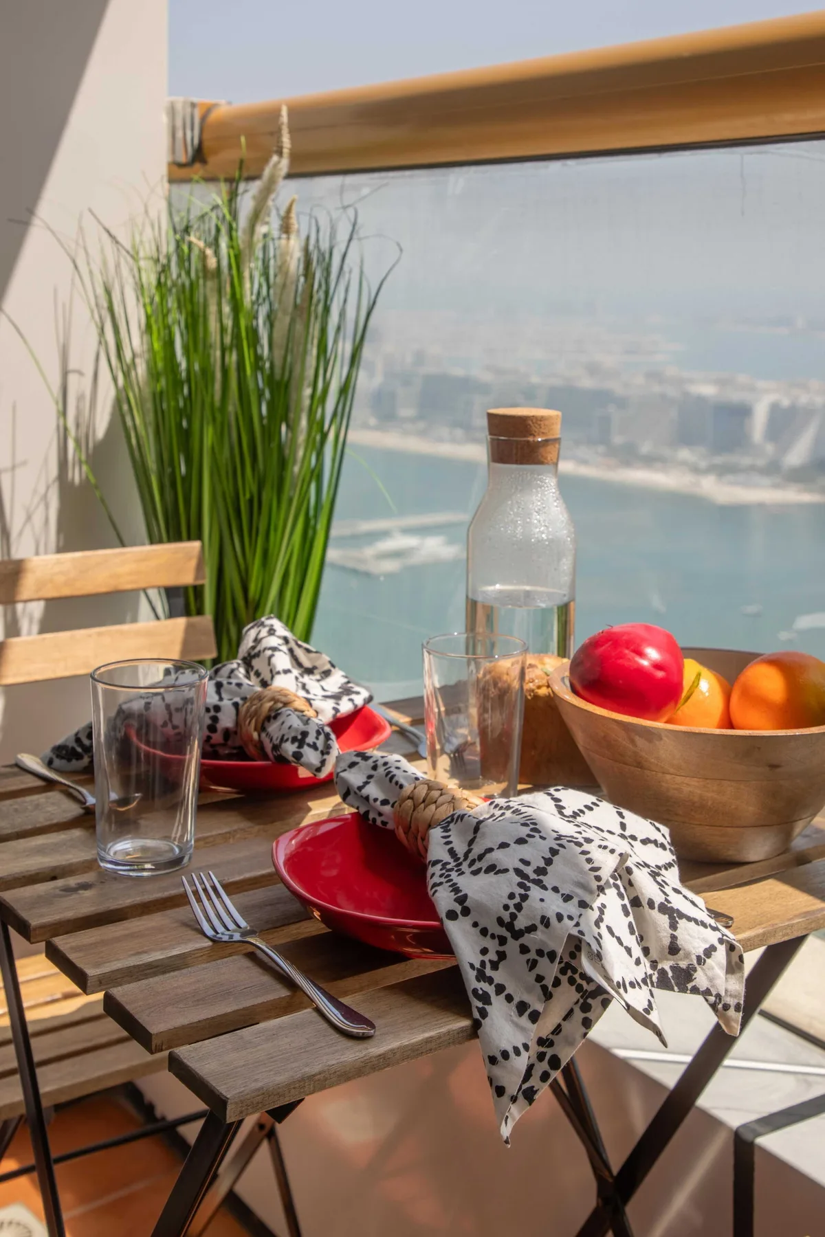 Una mesa de balcón soleada está preparada con platos, fruta y una botella de vidrio.