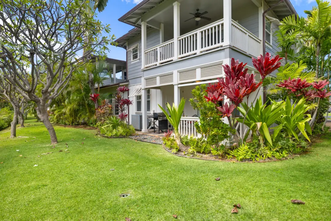 Una casa de dos pisos con balcón cubierto está junto a un césped verde y plantas tropicales.