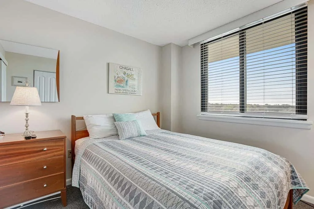 A bedroom with a made bed beside a wide window with blinds.
