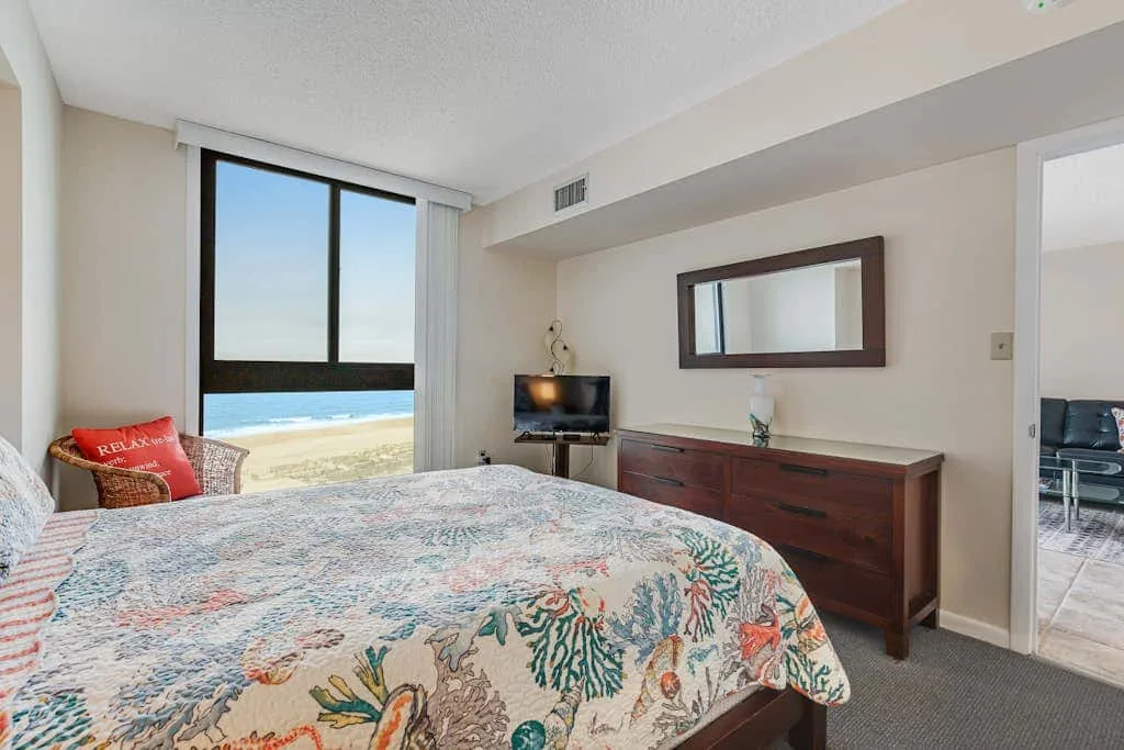 A bedroom with a patterned bedspread, dresser, and window overlooking the beach.
