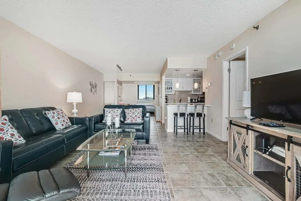 A living room with dark seating, a glass coffee table, and a kitchen beyond.