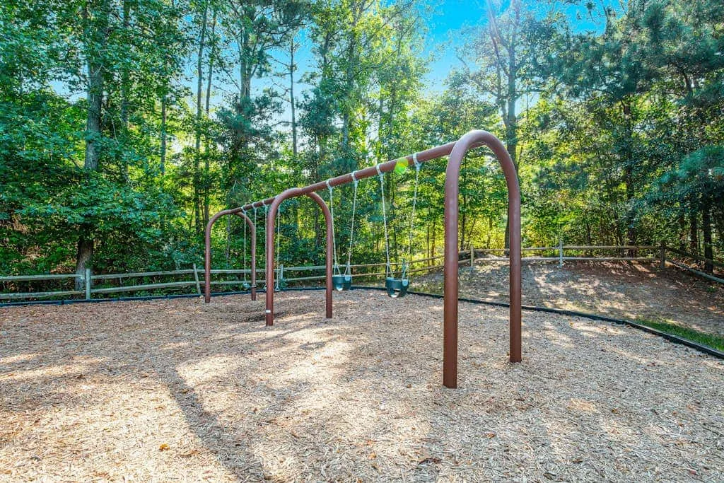 An outdoor playground swing set stands on a wood-chip surface among trees.