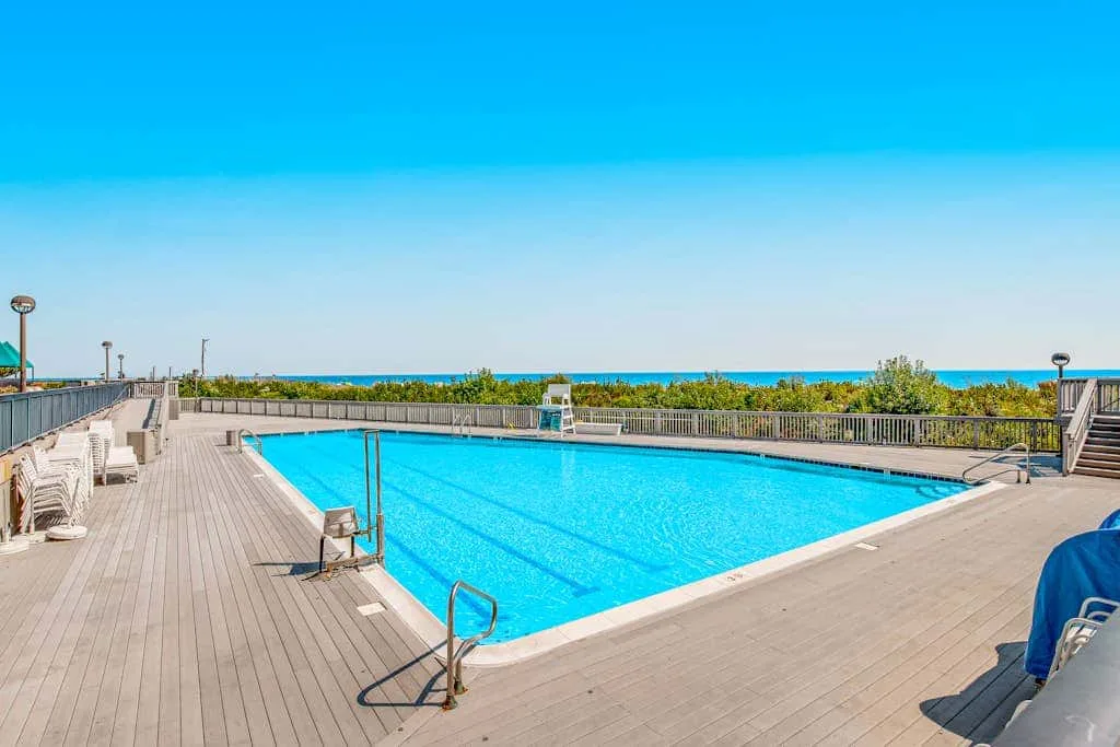 A large outdoor swimming pool sits on a sunny deck beside the ocean.