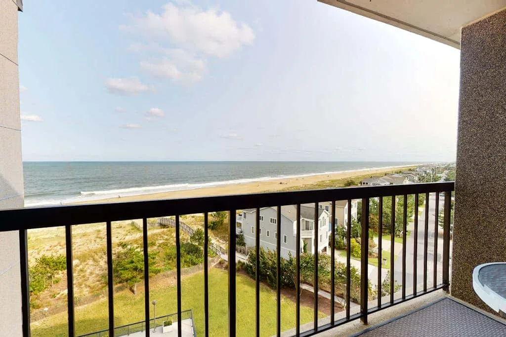 A balcony overlooks the beach and ocean, with a black railing in the foreground.