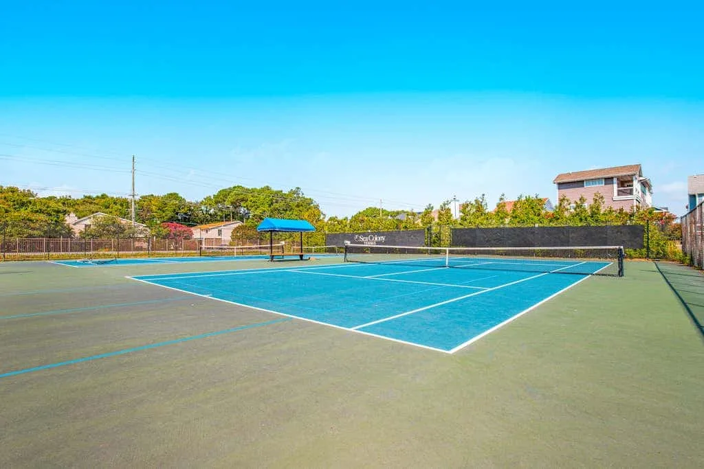 A blue tennis court with a net sits outdoors beneath a clear sky.