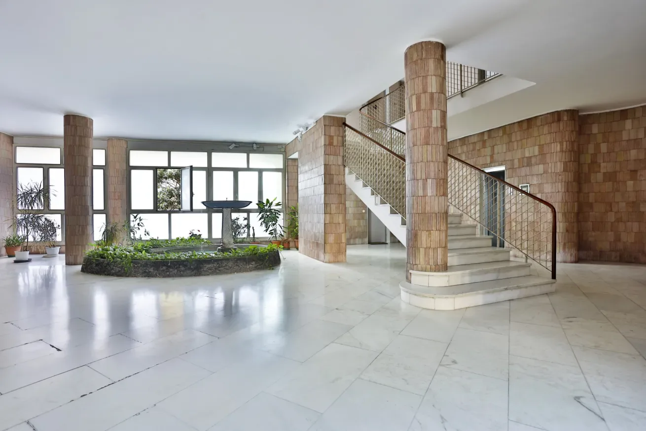 A spacious lobby with a tiled staircase and a planted area beneath large windows.