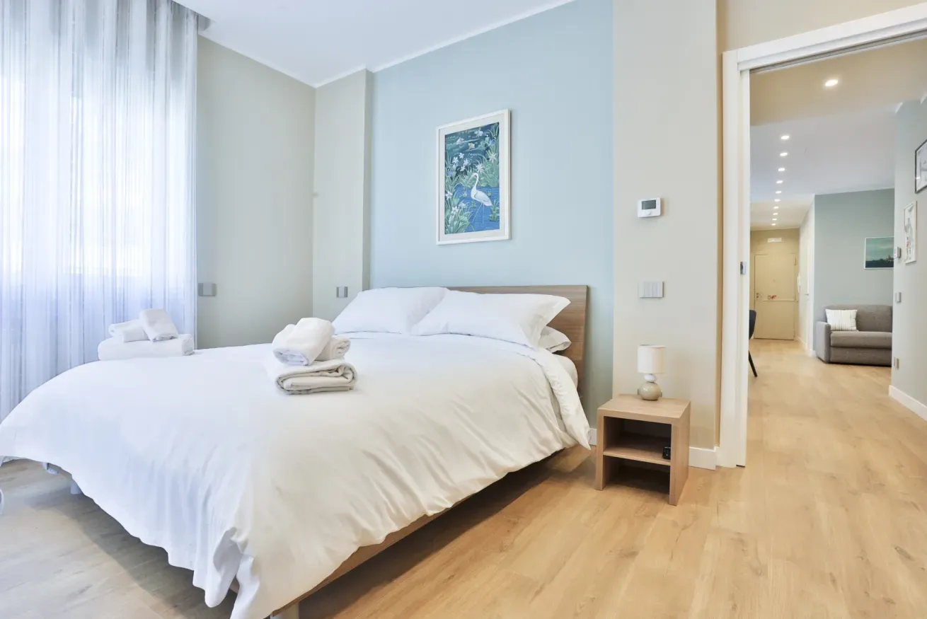 A bright bedroom with a neatly made bed and an open doorway to a sitting area.
