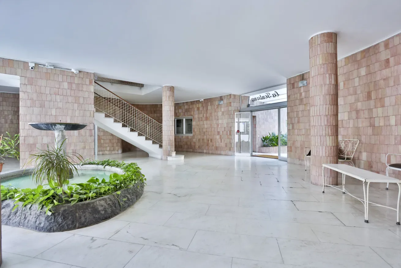 A spacious tiled interior lobby with a staircase, glass doors, and a planted fountain.