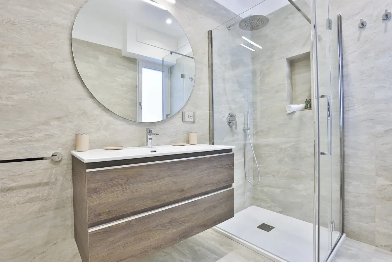 A modern bathroom with a wood-tone vanity, round mirror, and glass shower.