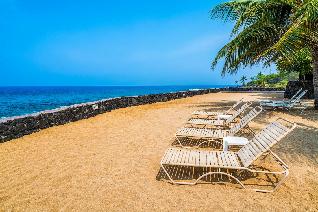 A sandy beachside area with lounge chairs beside a low stone wall and the ocean.