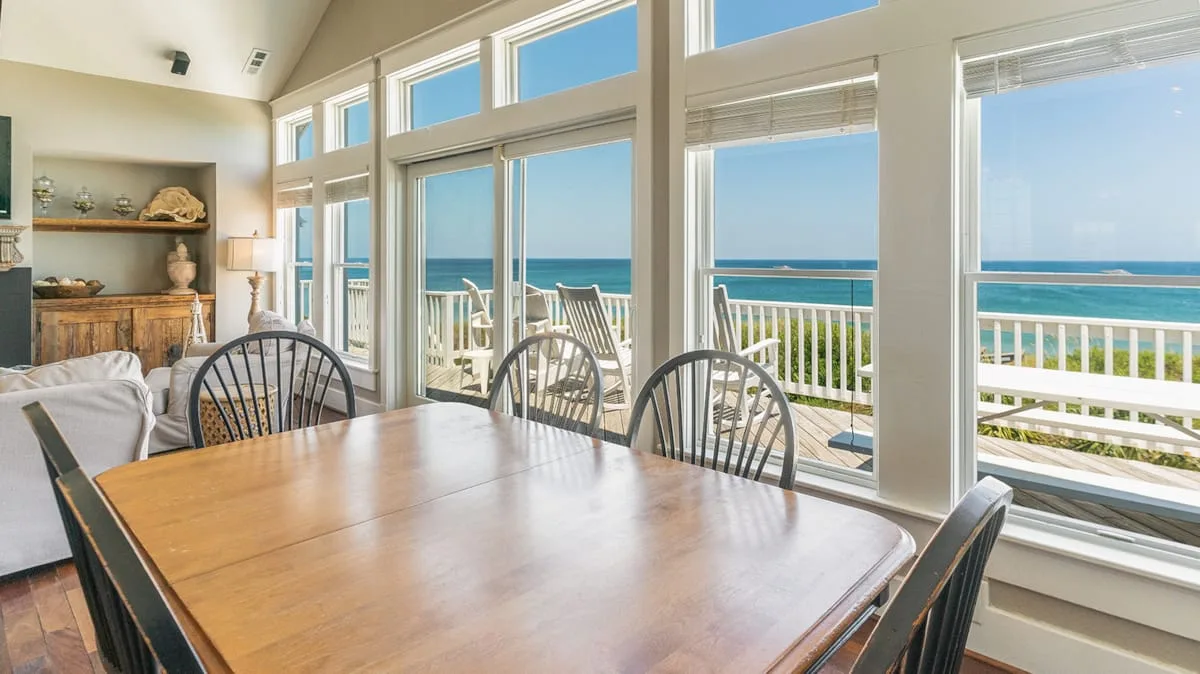 A dining area with a wooden table beside large windows overlooking the ocean.