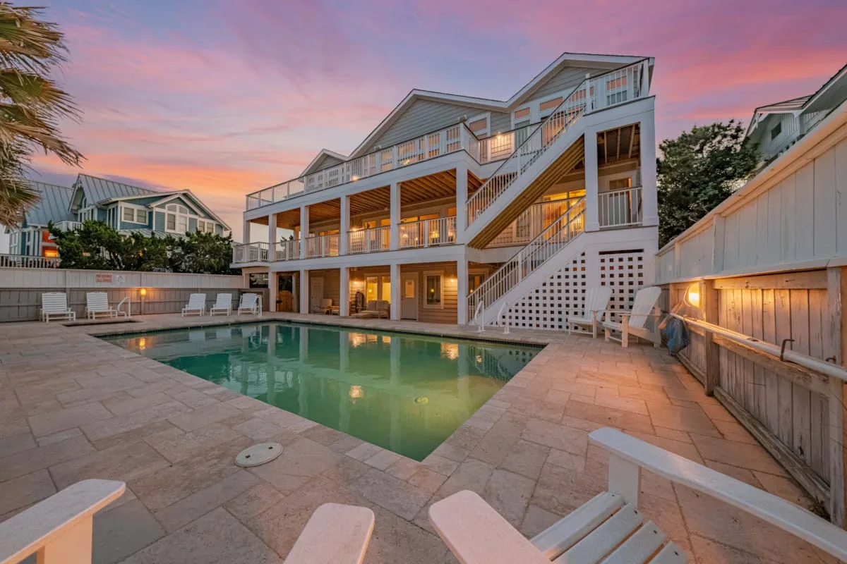 A swimming pool and paved patio sit beside a large house at sunset.