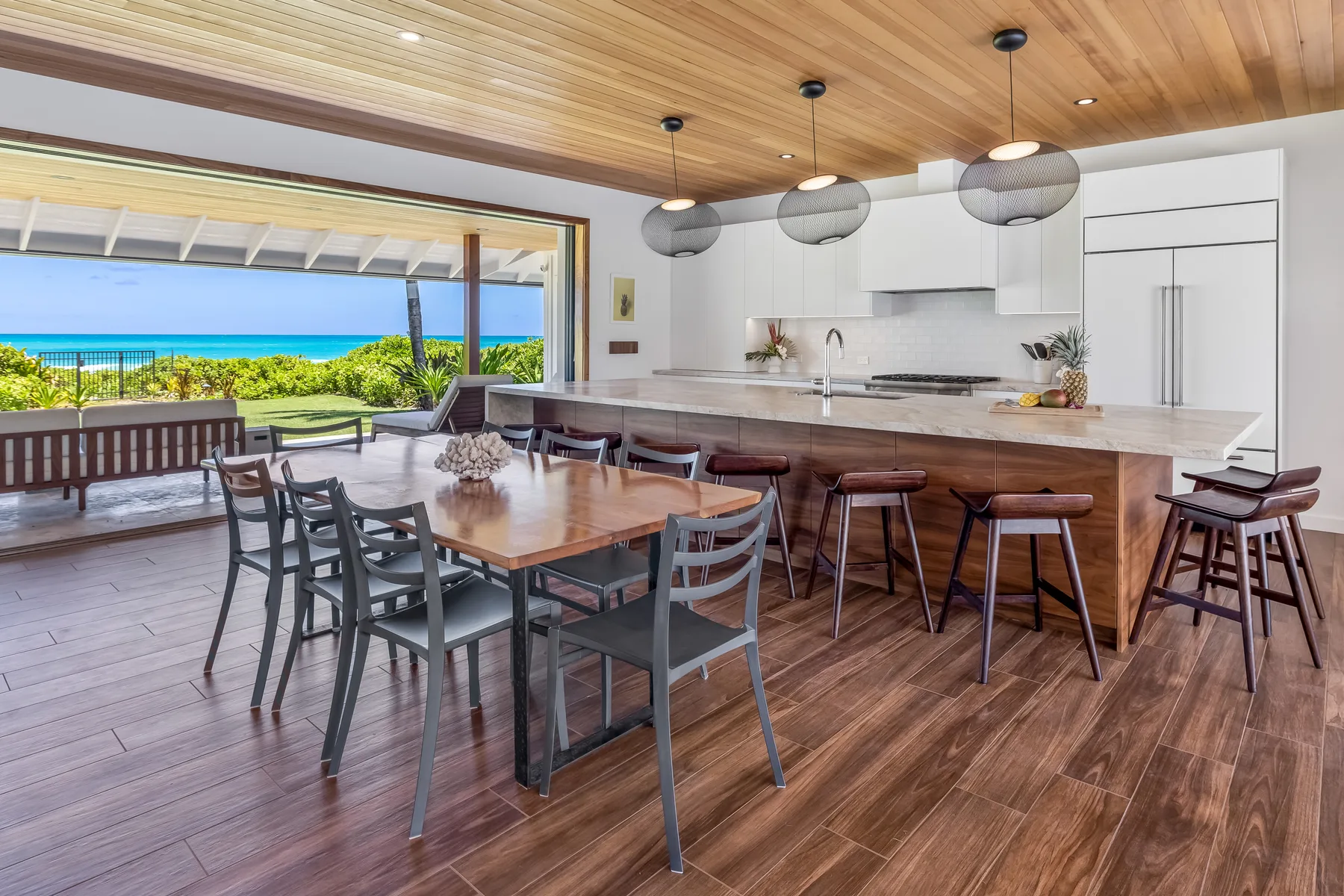 An open kitchen and dining area with a wooden table and a large window overlooking greenery and the ocean.