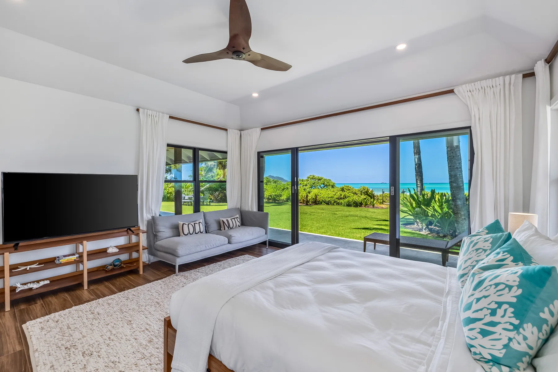 A bright bedroom with a bed, a seating area, and large windows overlooking a green lawn and ocean.