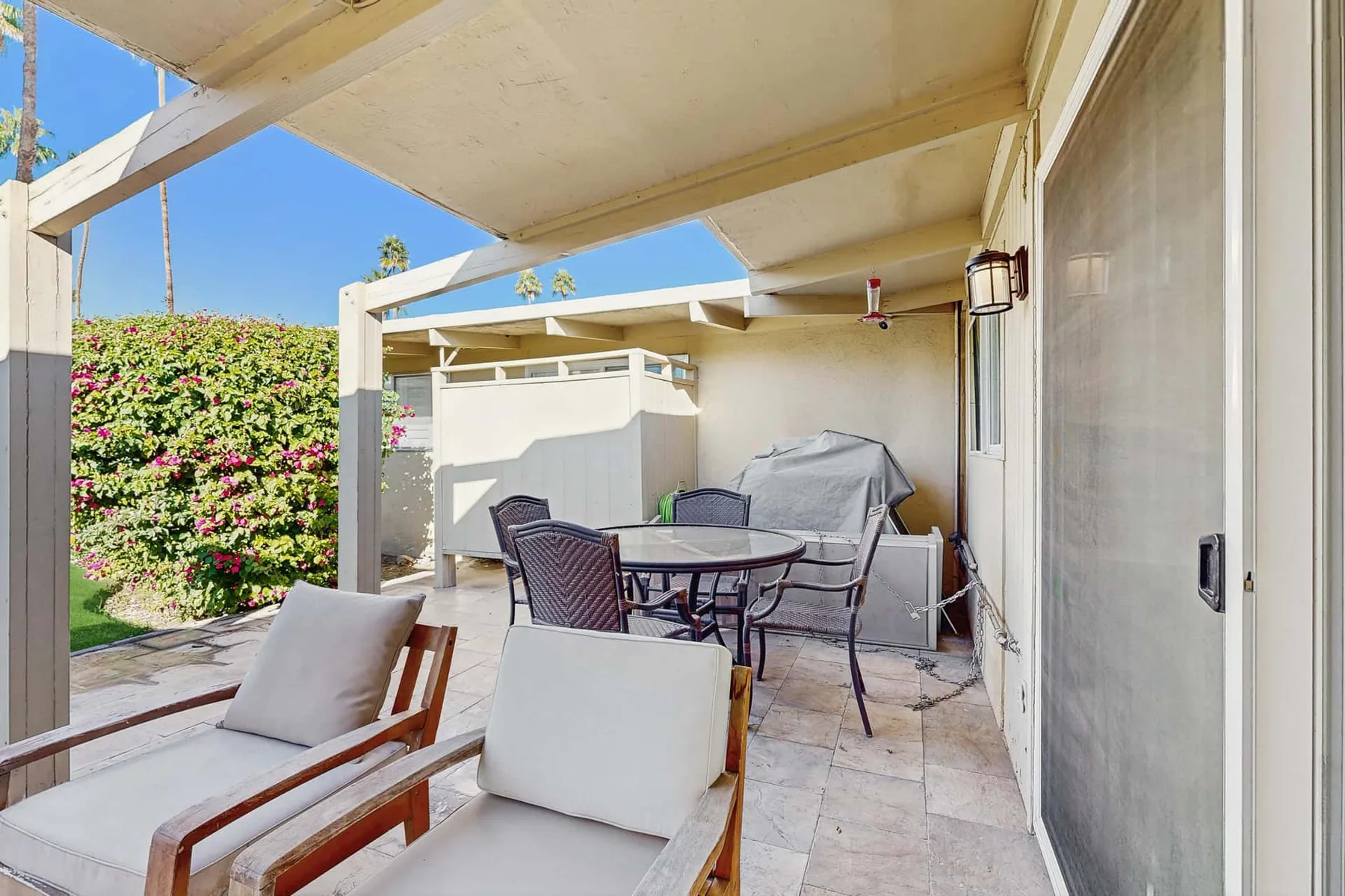 A covered patio has outdoor seating around a table beside a flowering hedge.