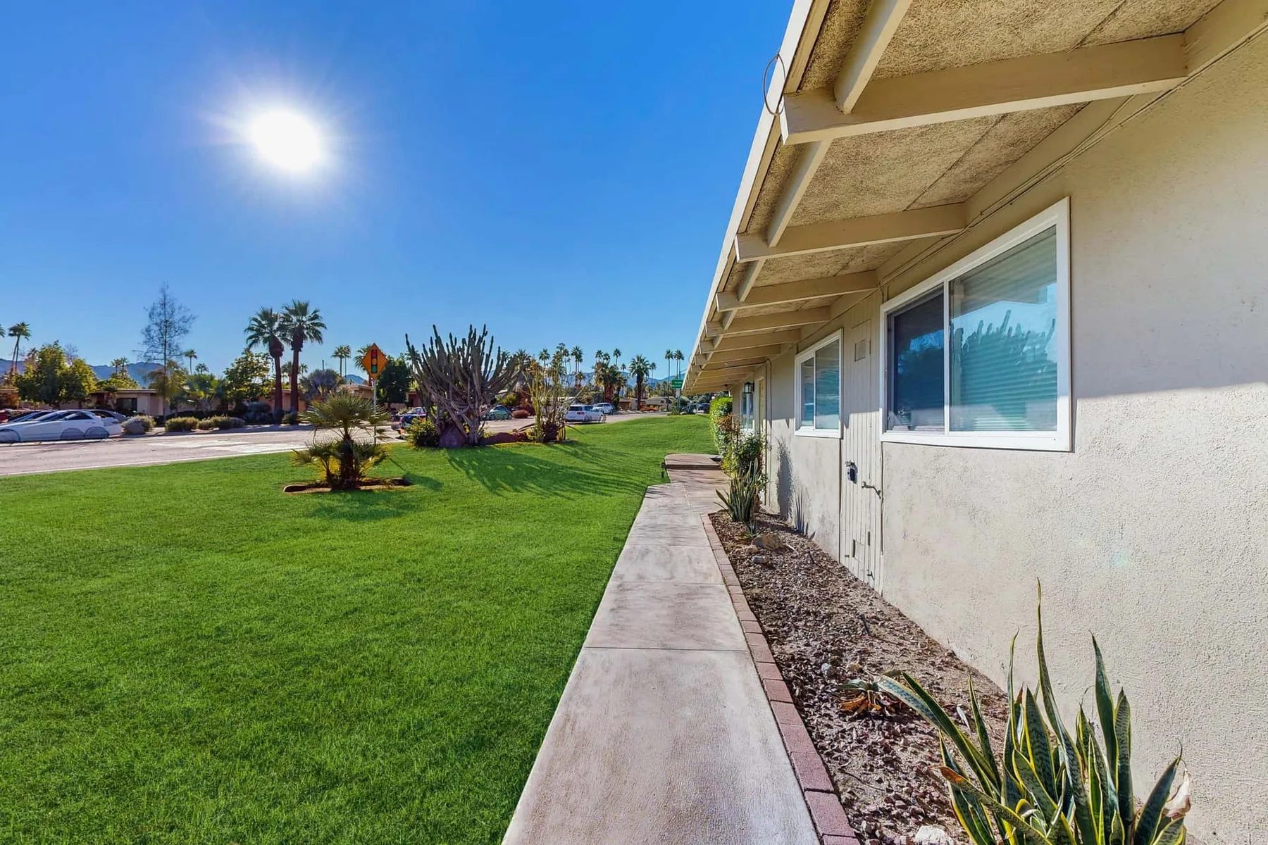 A sunny outdoor walkway runs beside a low building and landscaped lawn.