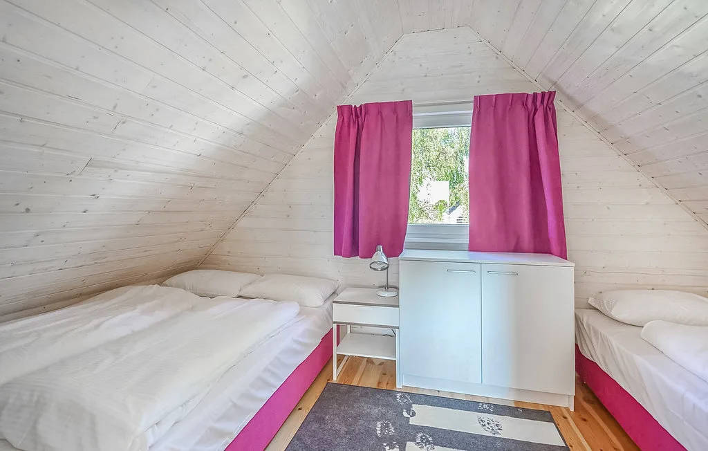 Un dormitorio abuhardillado luminoso con camas bajo un techo blanco inclinado y una ventana con cortinas.
