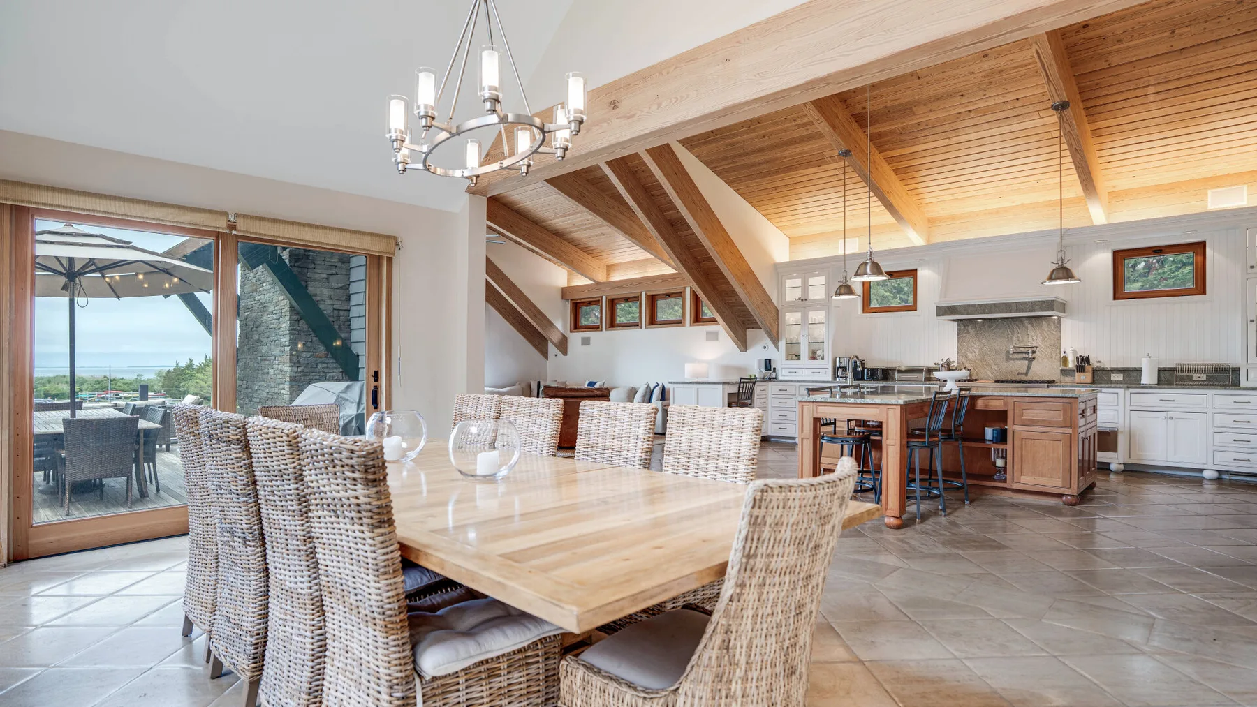 A dining area with a long table and an open kitchen beneath a wood-beamed ceiling.
