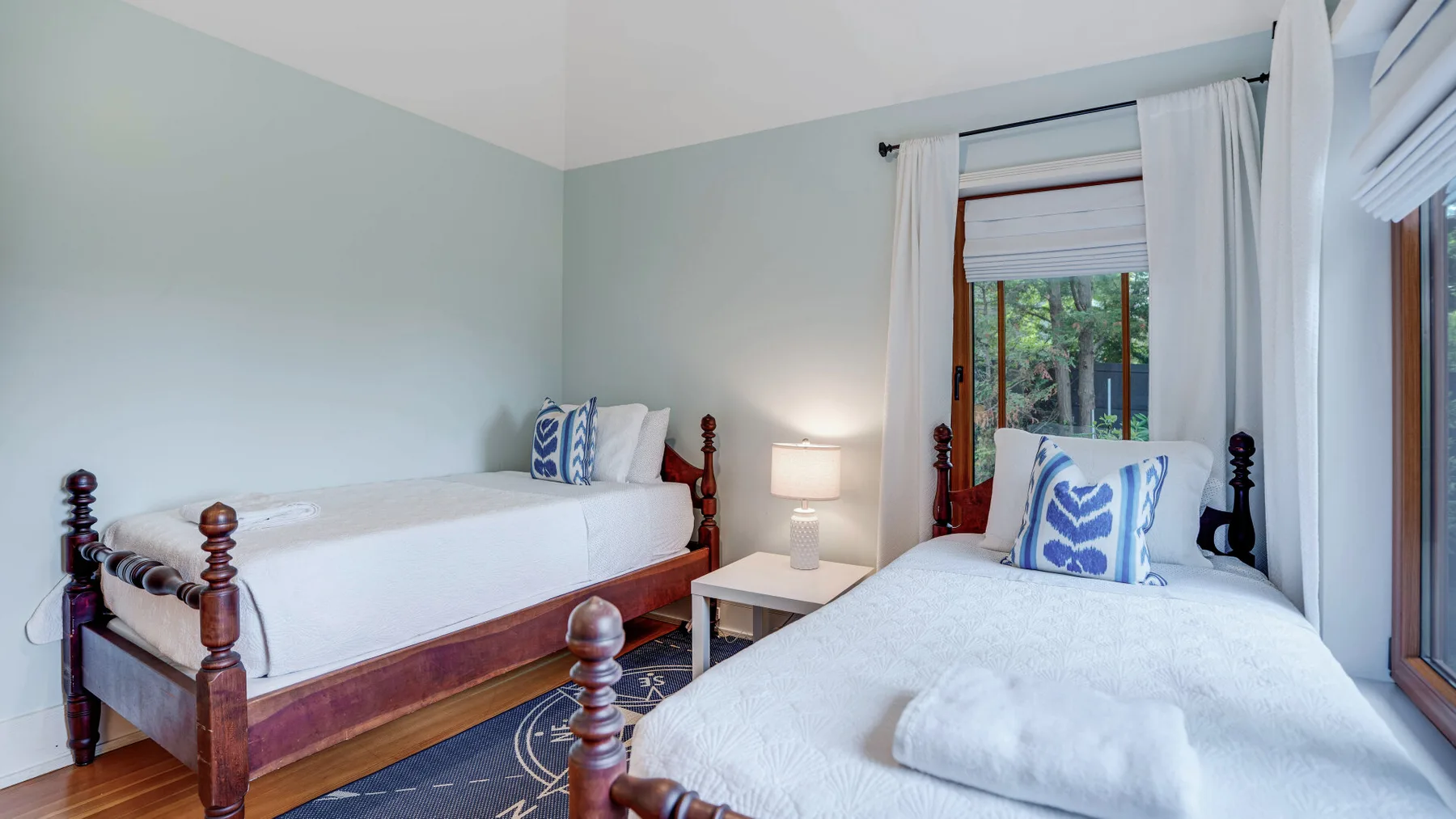 A bedroom with two single beds, white bedding, and windows overlooking greenery.