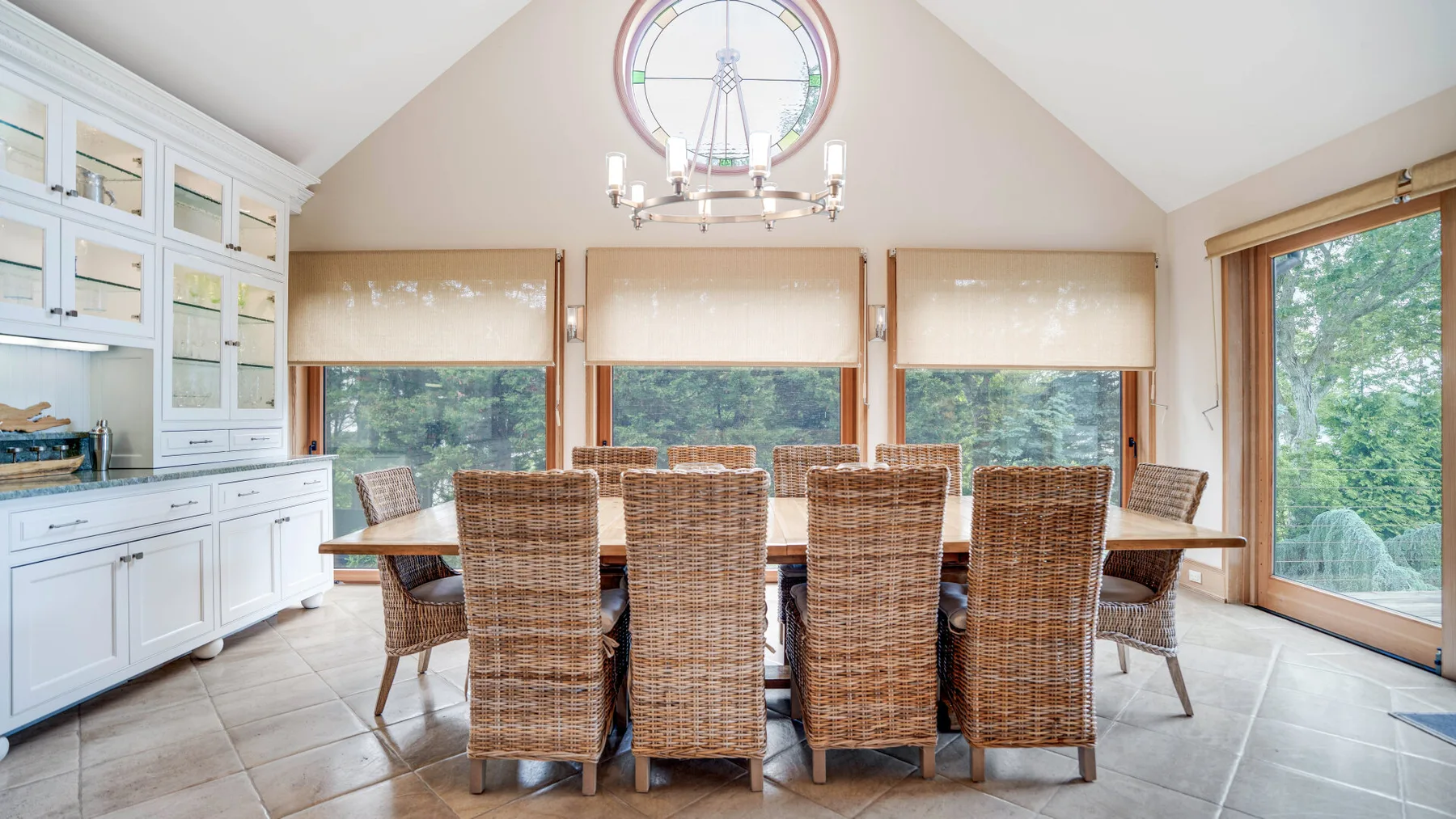A bright dining area with a long table and woven seating beneath a large round window.