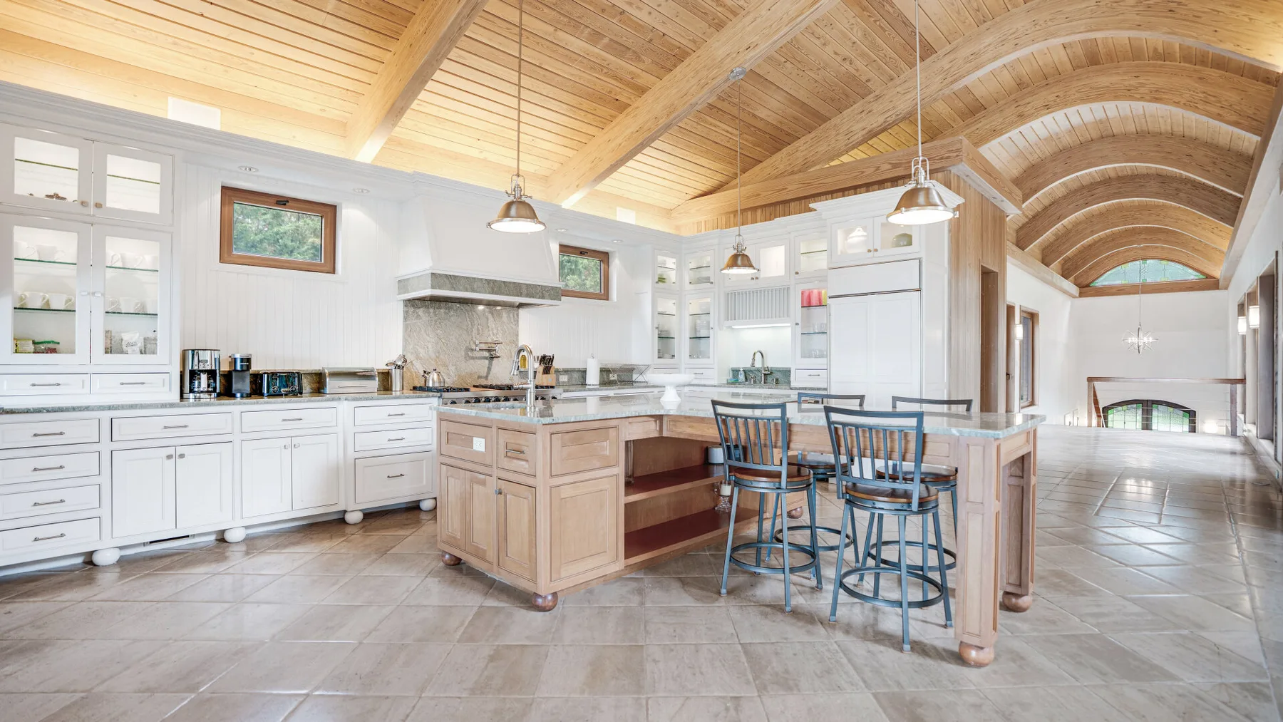 A spacious kitchen with a central island, dining stools, and a vaulted wood ceiling.