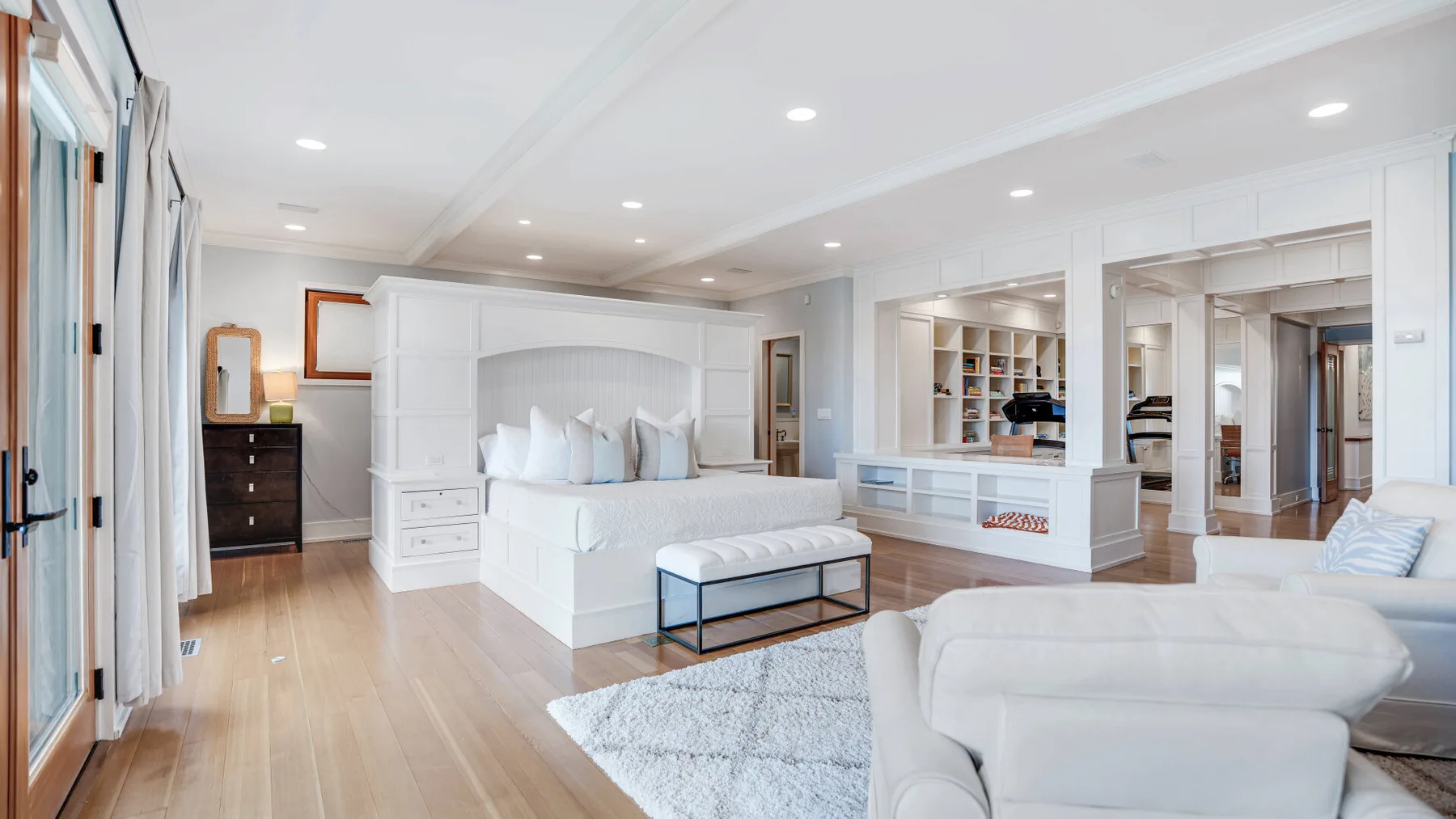 A bright bedroom with a large bed, a white seating area, and light wood flooring.