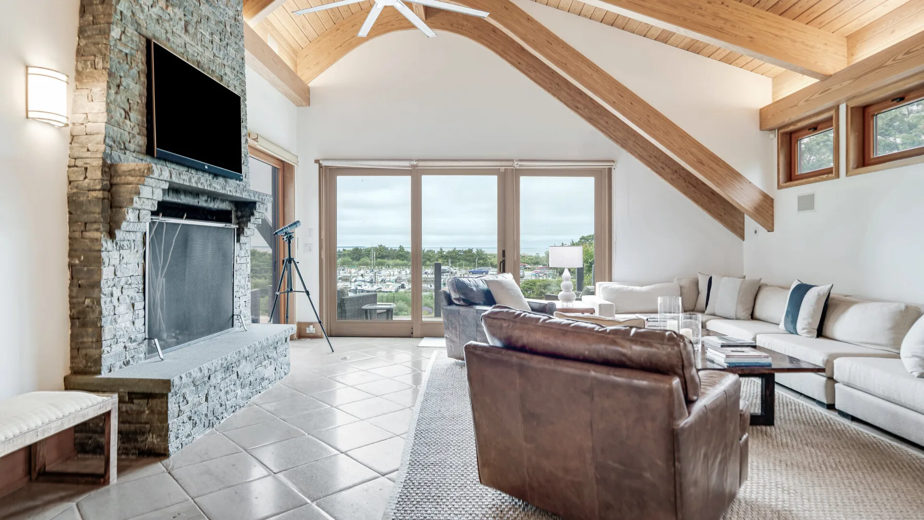 A bright living room with a stone fireplace, seating, and large windows.