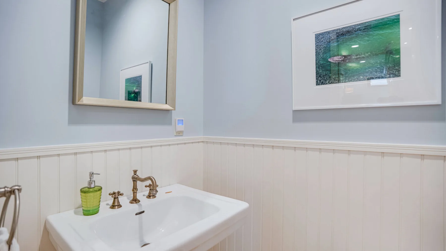 A bathroom sink sits beneath a mirror, with framed artwork on the wall.