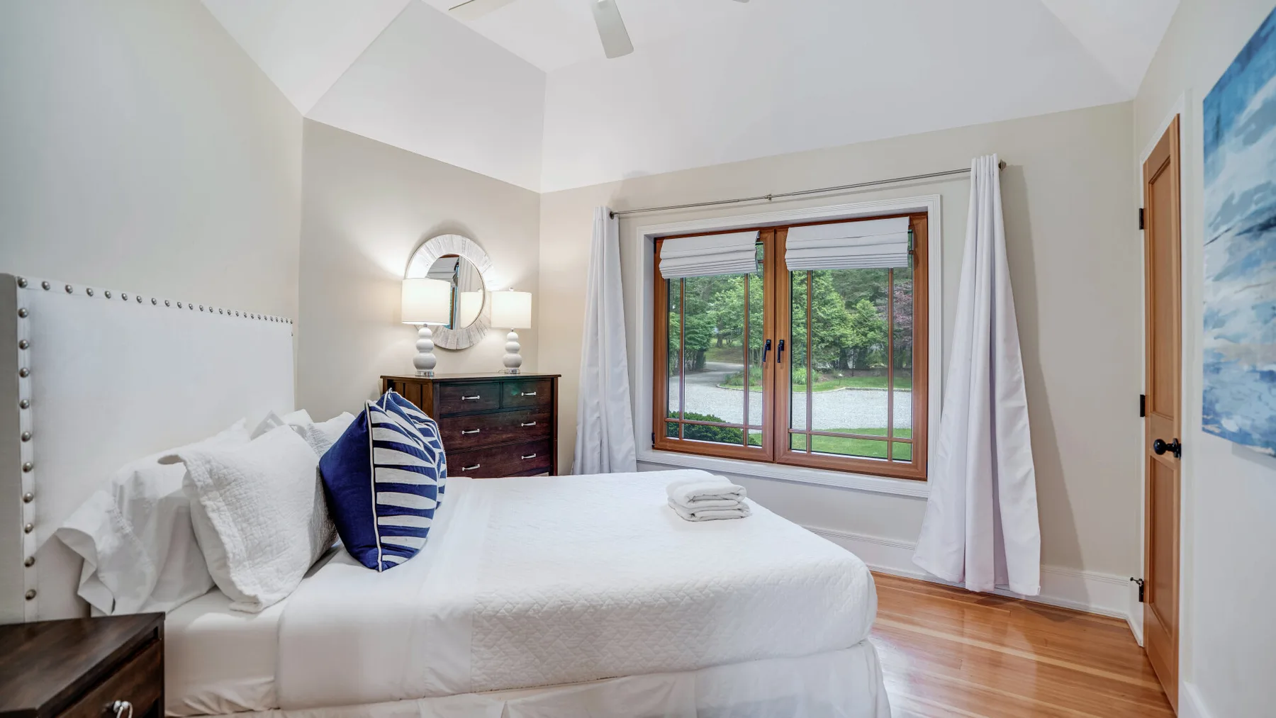 A bright bedroom with a neatly made bed beside a large window overlooking greenery.