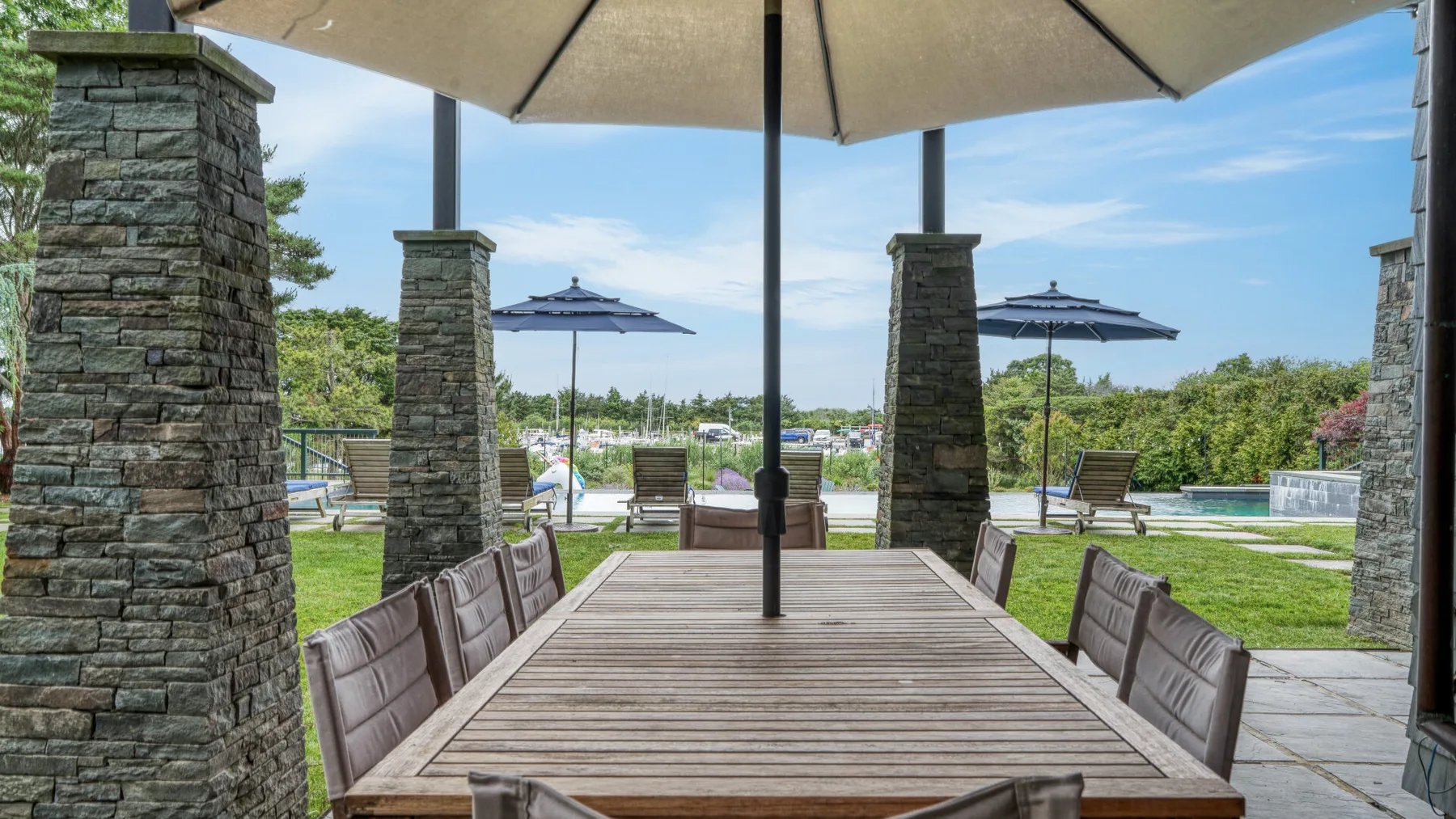 An outdoor dining area has a long table beneath a large umbrella, with seating around it.