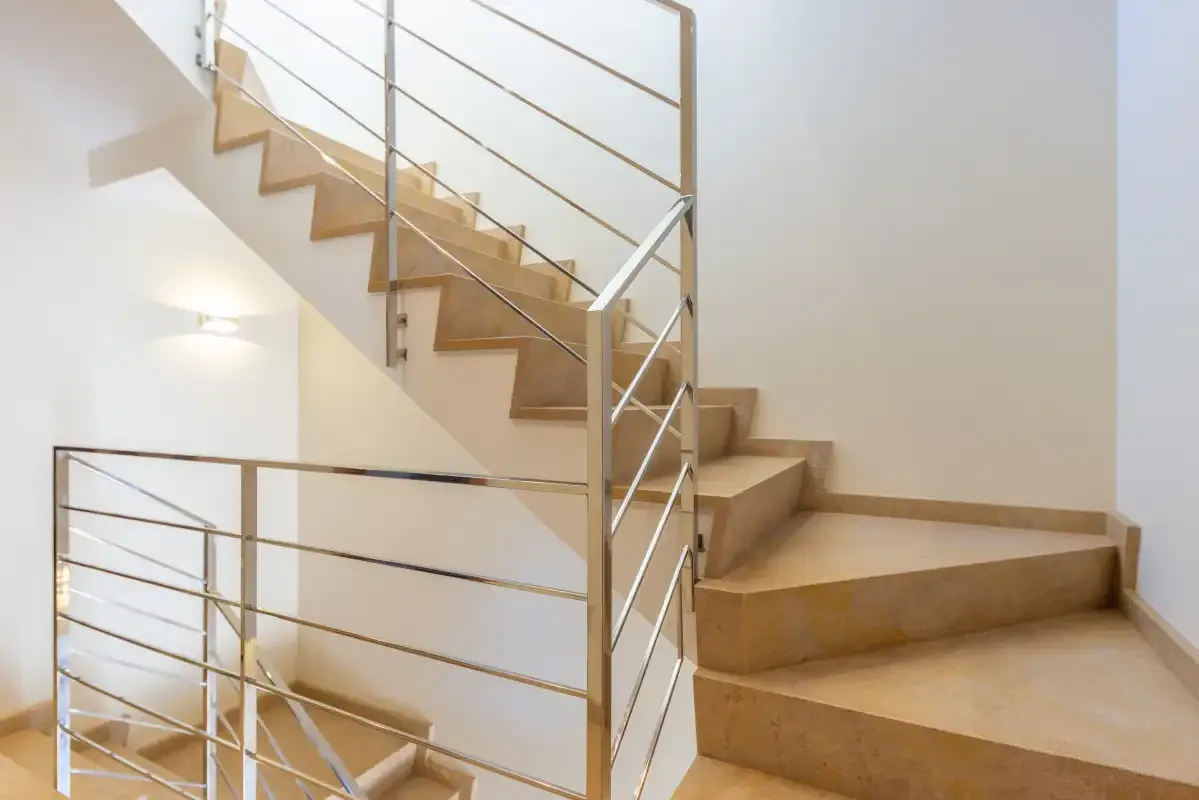 Una escalera con peldaños beige y barandillas metálicas gira junto a una pared blanca.