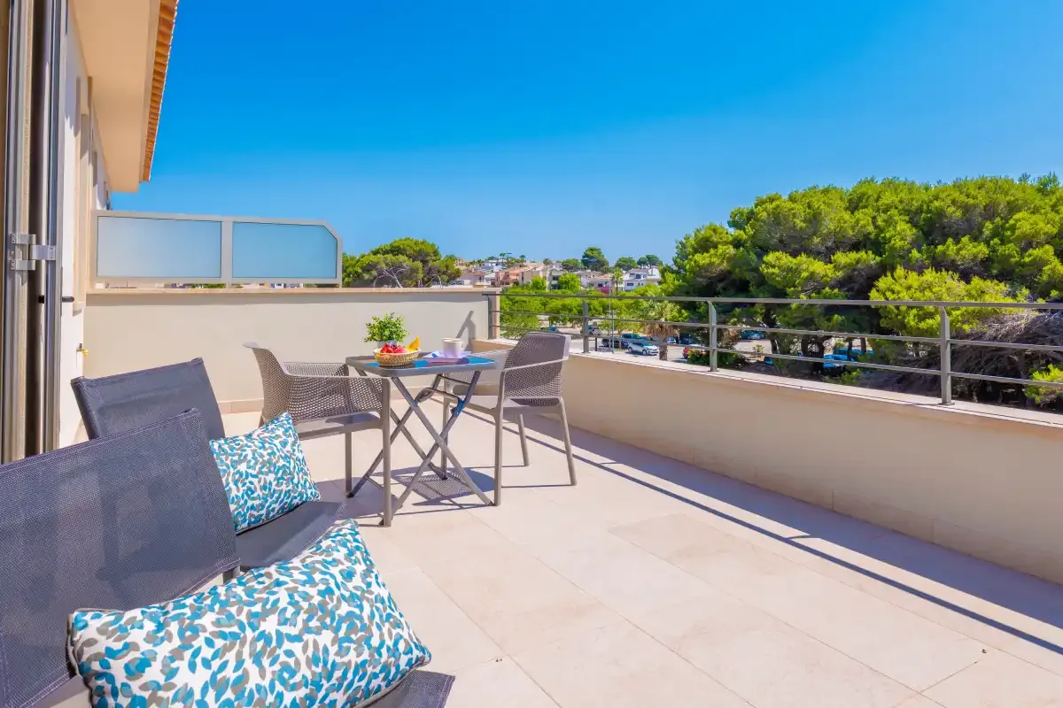 Una terraza soleada tiene asientos y una mesa al aire libre, con árboles y edificios cercanos a la vista.