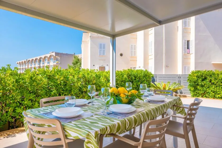 An outdoor dining table is set beneath a covered patio beside a green hedge.