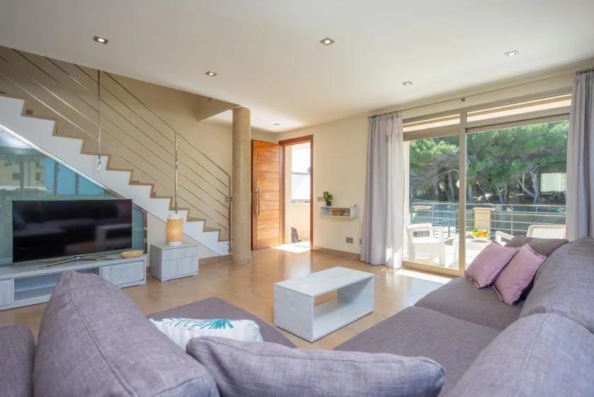 Una luminosa sala de estar con asientos, un televisor y puertas de vidrio hacia un patio.
