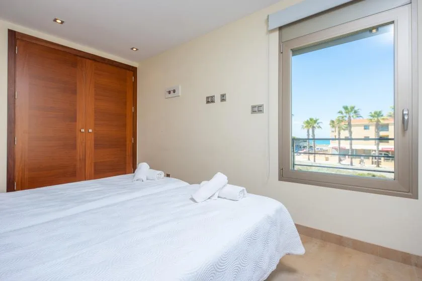 Un dormitorio con una cama bien tendida junto a una ventana amplia con vistas a palmeras y edificios.