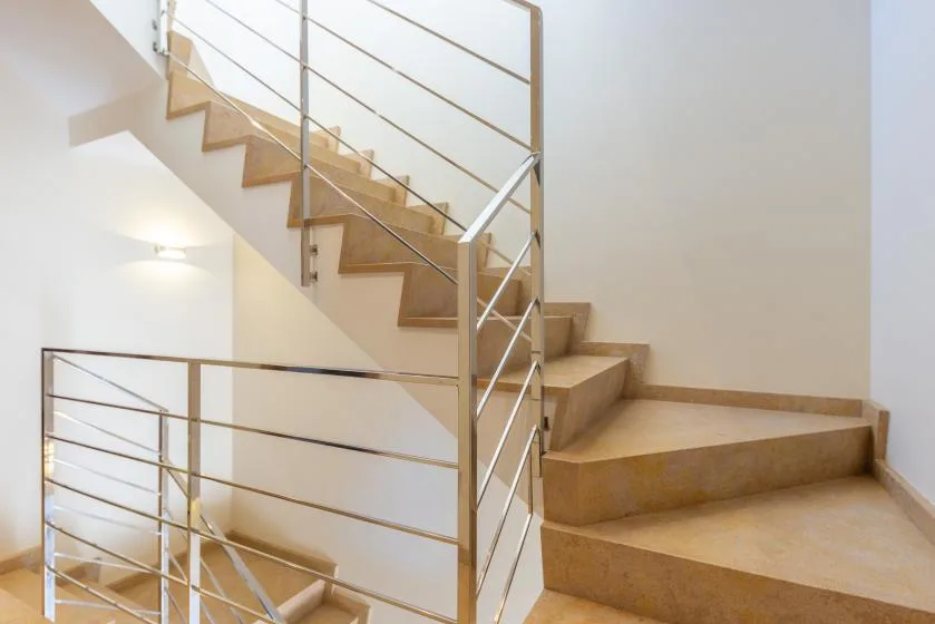 Una escalera de peldaños color tostado y barandillas metálicas asciende por un interior luminoso.