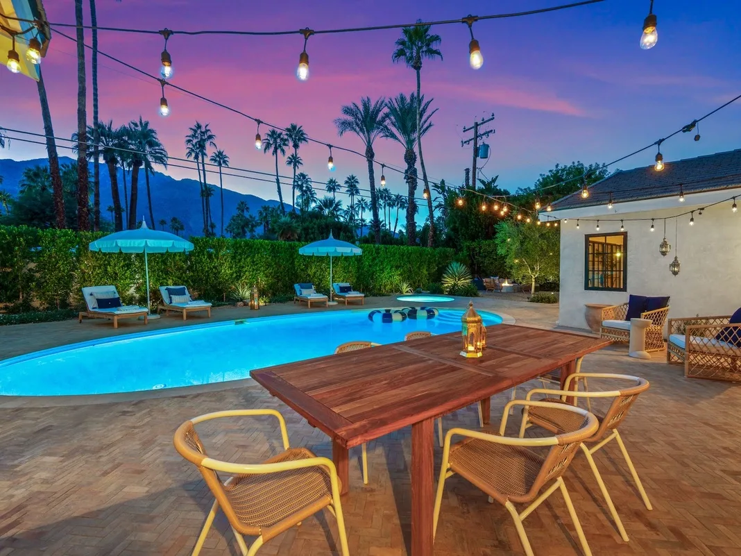 Un patio junto a una piscina al aire libre, con asientos y luces colgantes al anochecer.
