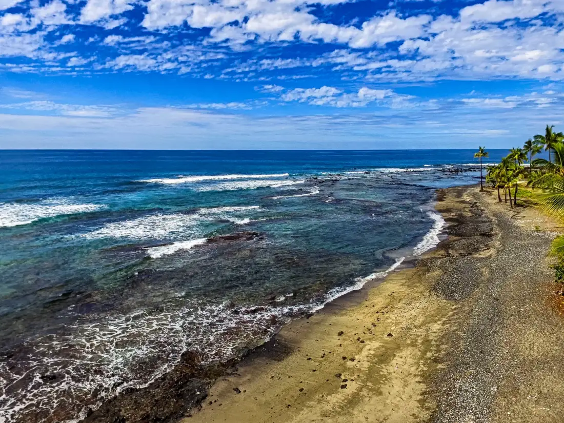 Una costa tropical con olas rompiendo sobre una orilla rocosa bajo un cielo azul y nublado.