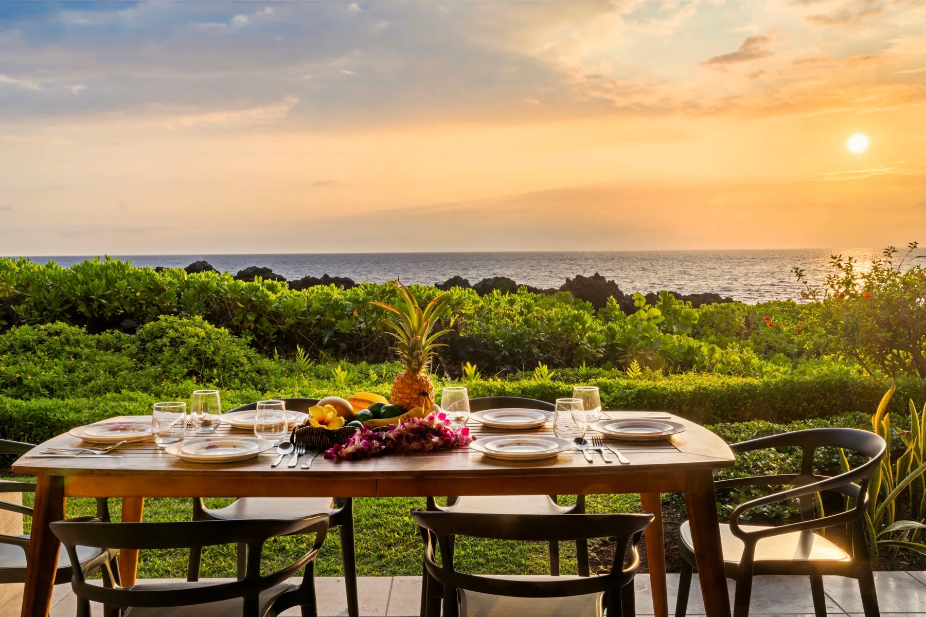 Una mesa de comedor al aire libre está preparada junto a un jardín verde con vistas al mar.
