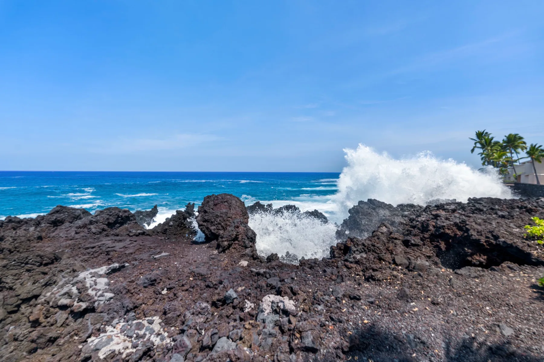 Las olas rompen contra una costa rocosa oscura bajo un cielo azul despejado.
