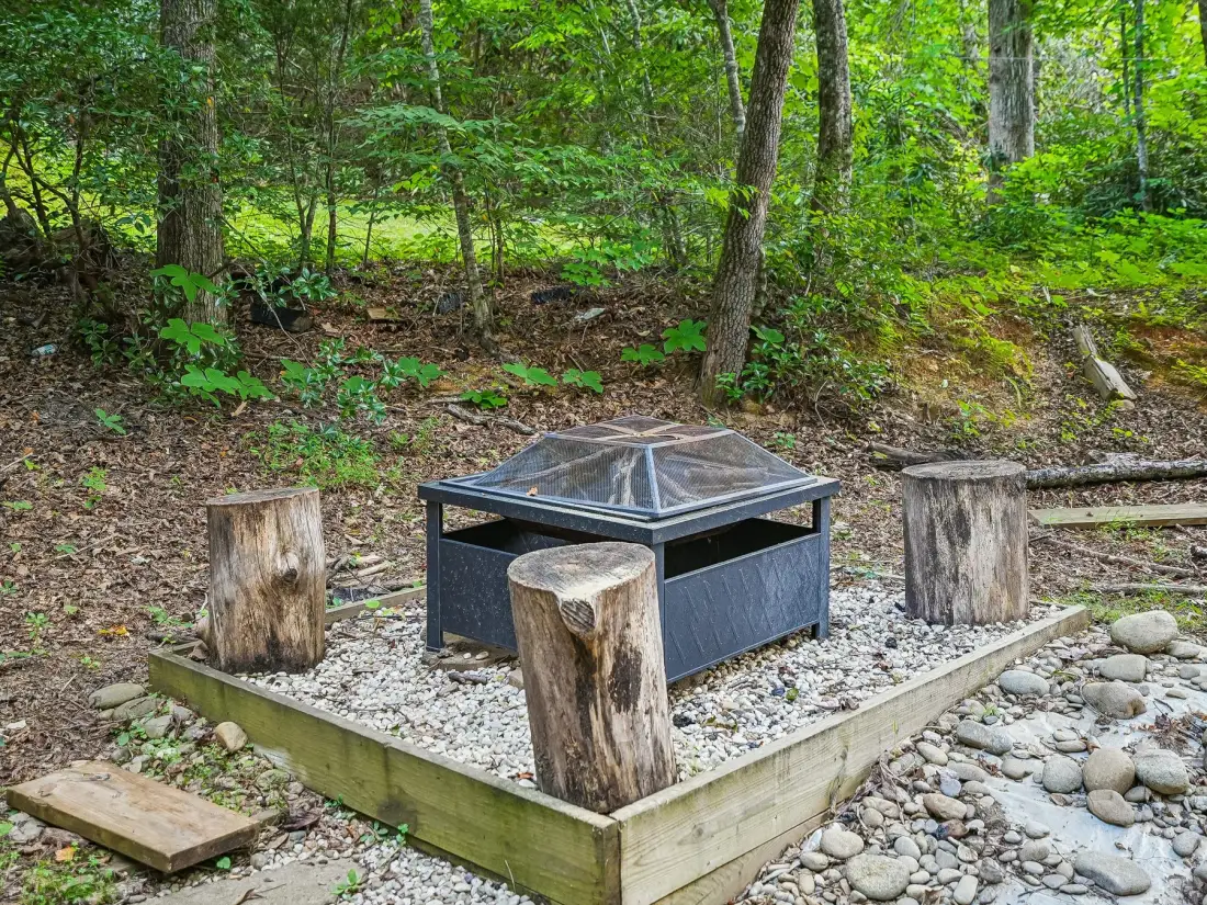 A fire pit sits in a gravel clearing surrounded by trees and cut-log seating.