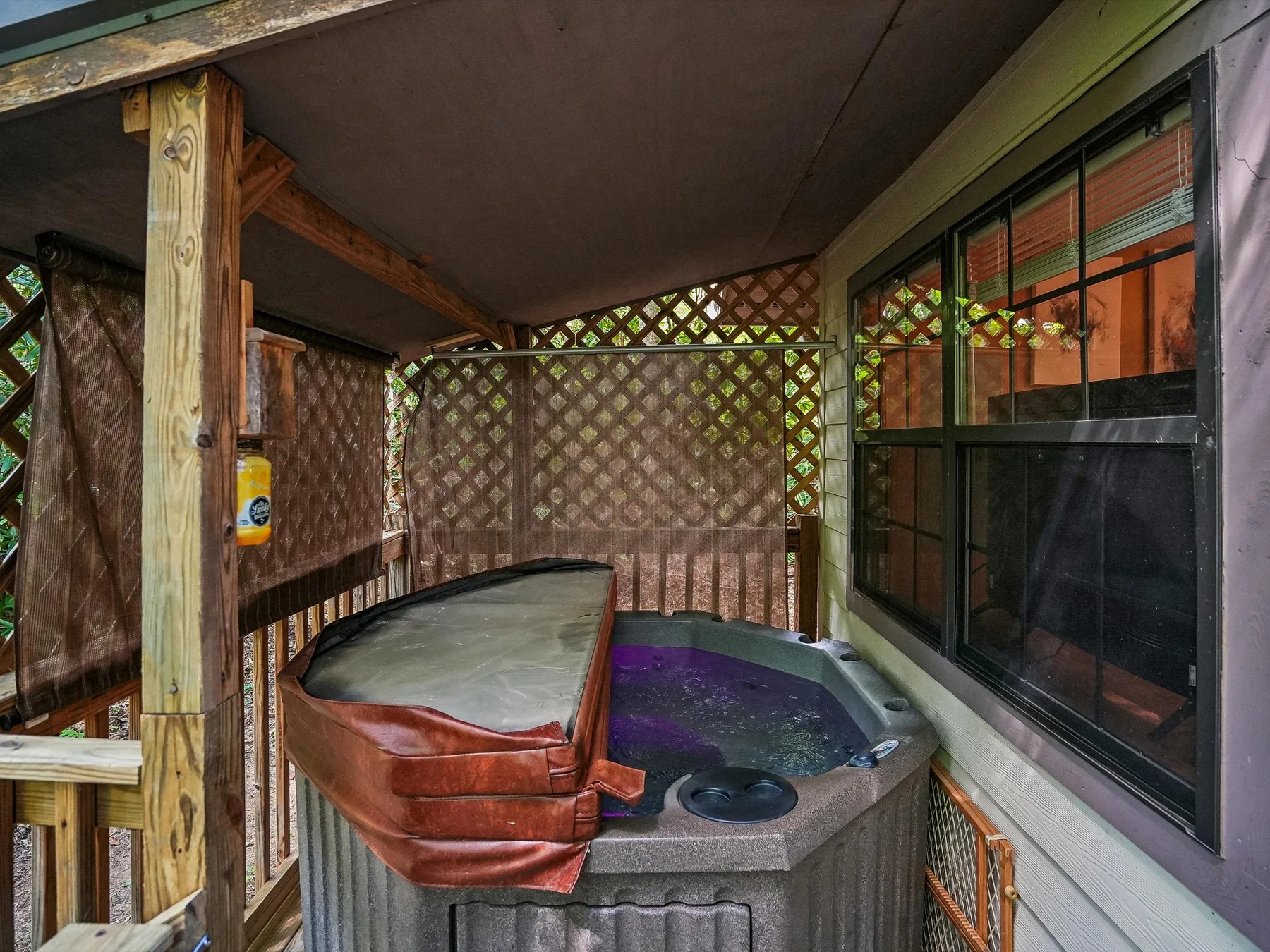 A covered outdoor hot tub sits beside a large window and lattice fencing.